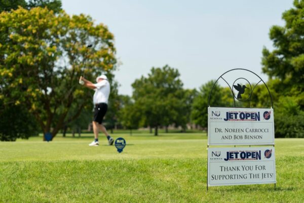 A golfer takes a swing on the golf course. A Jet Open sign reads "Dr. Noreen Carrocci and Bob Benson, thank you for supporting the Jets!"