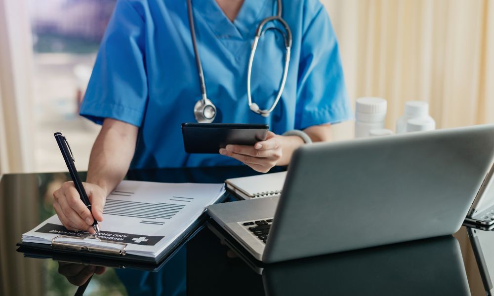 A doctor sits with a laptop, documentation pad, and tablet.