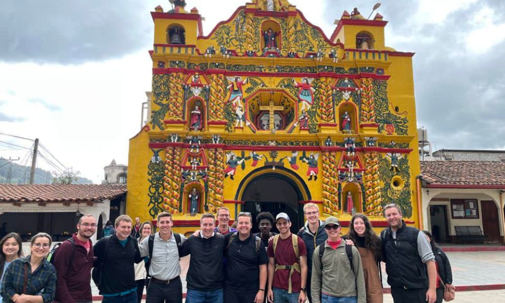 Newman students and seminarians from the Diocese of Wichita pose in front of the colorful facade of the church on the plaza at San Andres Xecul in Guatemala. Rev. Sam Brand is pictured at the far right.
