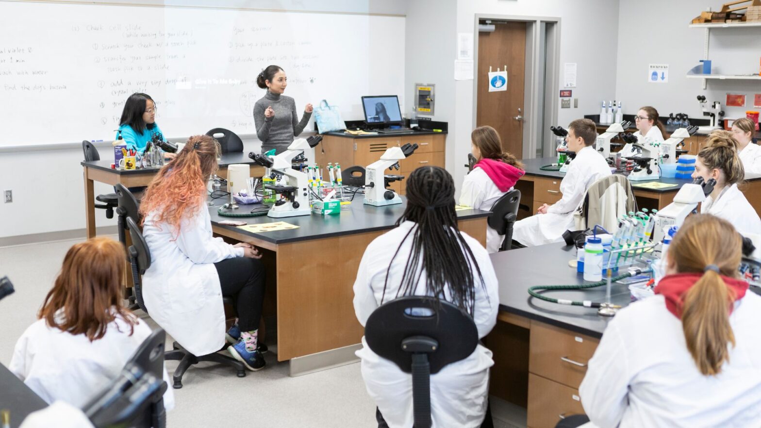 Newman professor Tomoko Bell teaches high school students wearing white lab coats in the Bishop Gerber Science Center.
