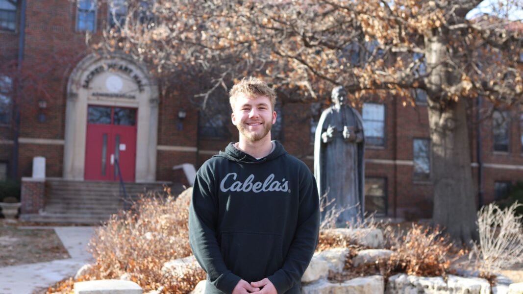Gilchrist, wearing a Cabela's sweatshirt, stands outside of Sacred Heart Hall on the Newman University campus.