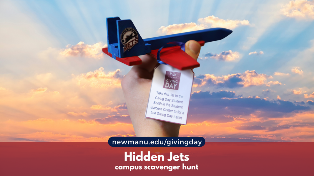 A hand holds up a wooden red and blue Newman University Jet with a Kansas sunset in the background. 