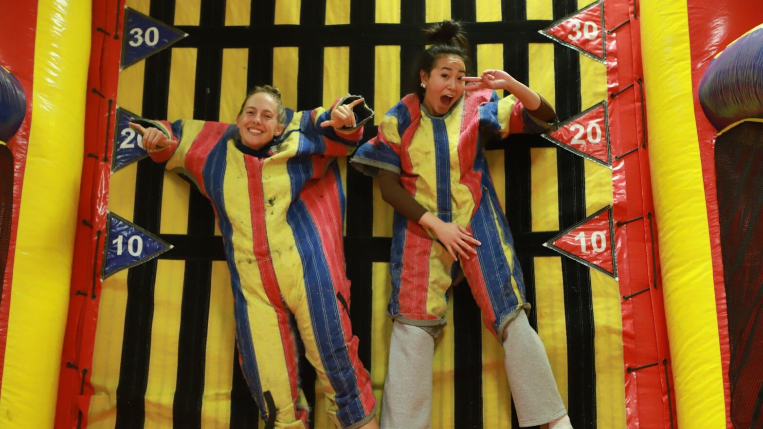 Two students wear yellow, red and blue-striped VELCRO outfits as they stick to an inflatable VELCRO wall.