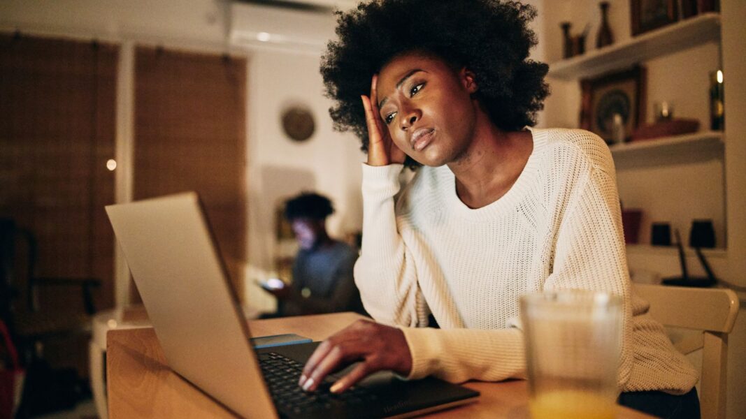 A Black woman looks frustrated with a hand on her head as she works on her computer