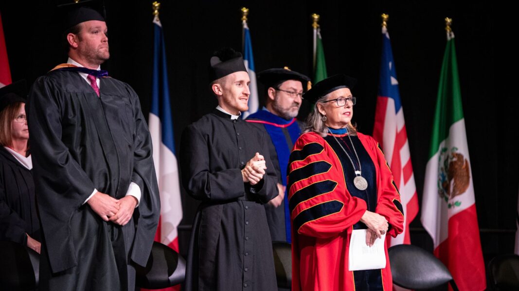 Father Adam Grelinger (second from the left) folds his hands on stage at the 2024 Newman commencement ceremony.