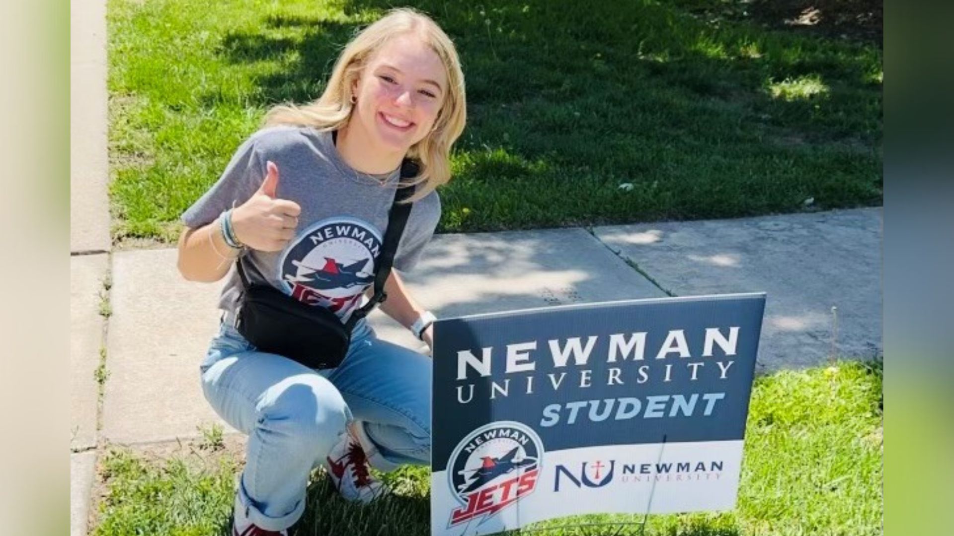 A smiling Newman student gives a thumbs up next to her "Newman University student" sign in her front yard.