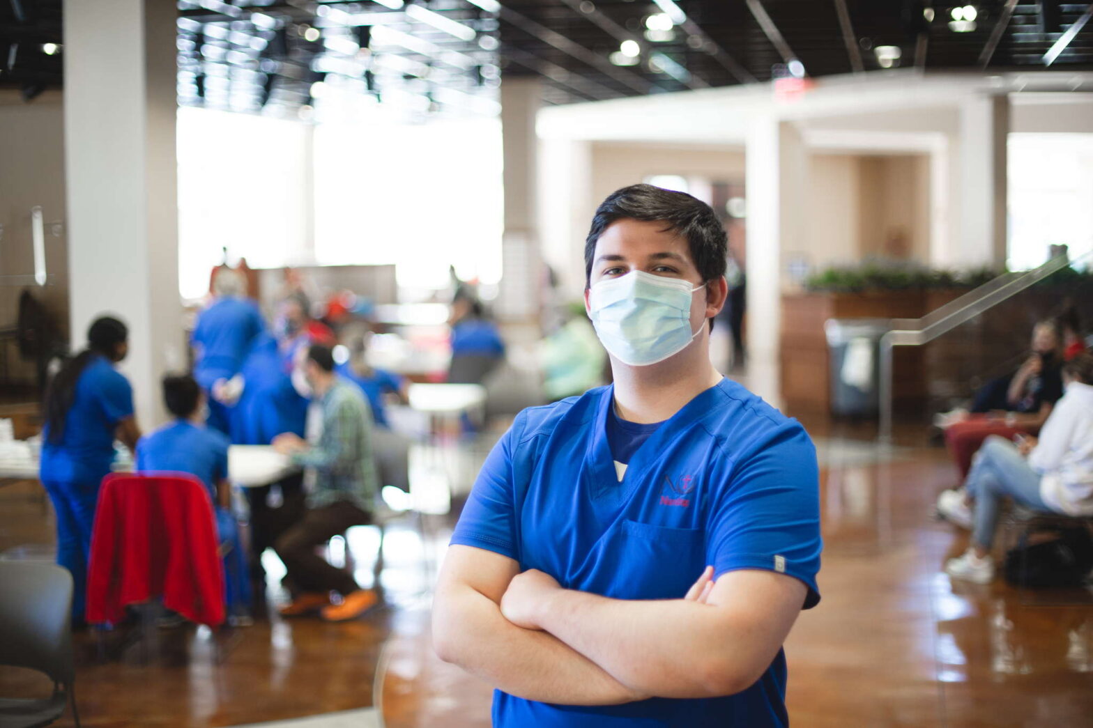 A male student wears a mask, scrubs, and crosses his arms during a health care professions event at Newman.