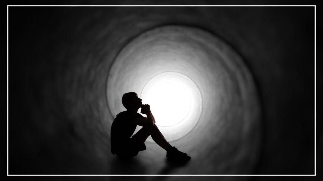 A person sits in darkness with hands folded in prayer with a circle of light behind them.