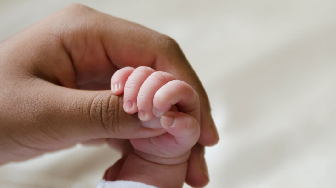 A baby's hand holds onto the thumb of a woman