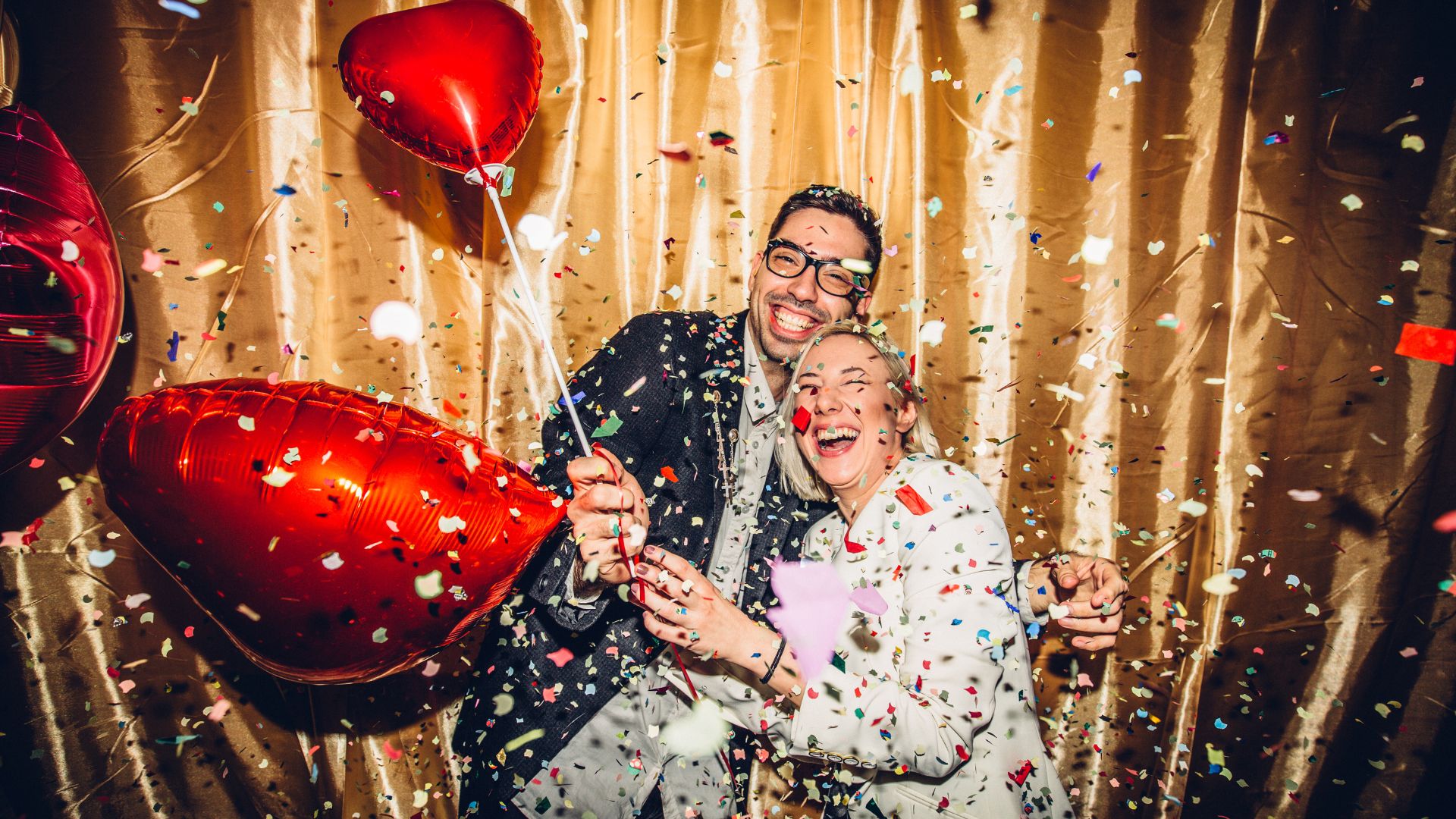 A man and woman smile as confetti and heart-shaped balloons make up the photo booth