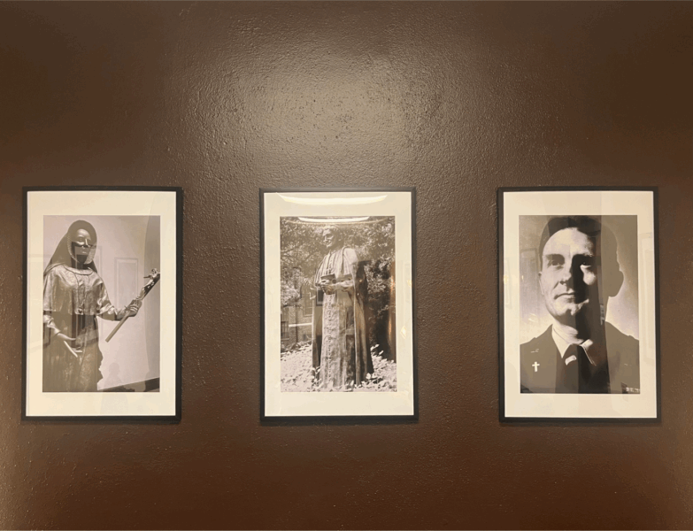 Three large framed photos on a deep brown wall, picturing St. Maria De Mattias, St. John Henry Newman, and Father Emil Kapaun