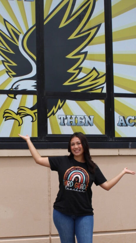 Pictures Yazmin in a third grade teacher shirt, in front of White Elementary School mural